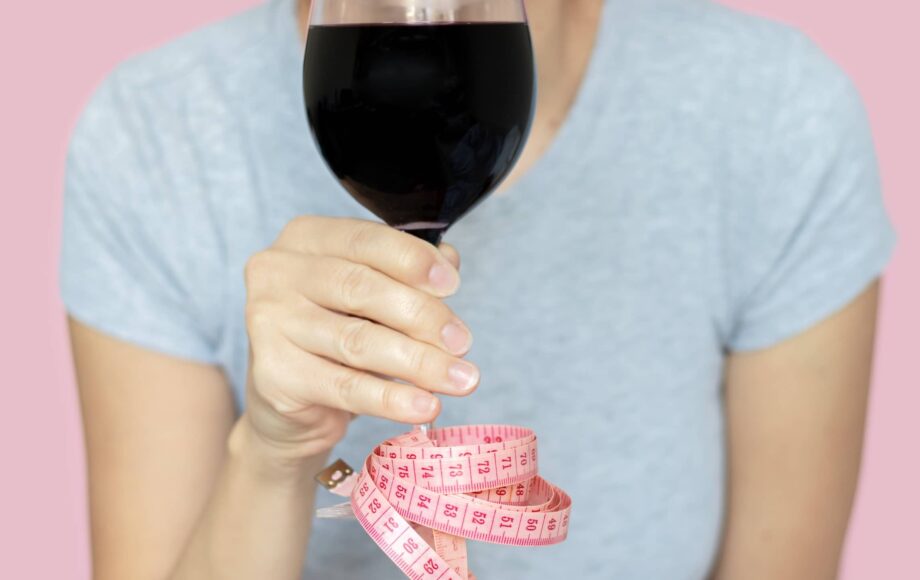 slim woman holding glass with red wine in hand measure tape attached by foot base